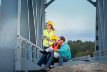 Railway and road engineers are standing in orange and white helmets and signal vests with documents against the background of the metal structure of the bridge.