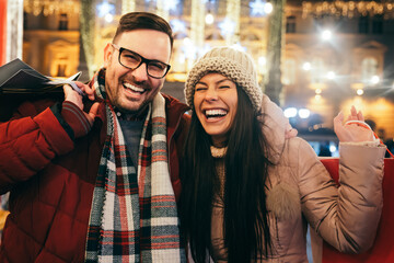 Cheerful happy couple doing Christmas shopping standing on decorated street. Sale people concept - obrazy, fototapety, plakaty