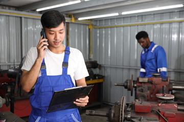 Multiethnic technicain mechanic using metal lathe machine operate polishing car disc brake with smart mobile phone and clipboard at garage. Maintenance automotive and inspecting vehicle part concept