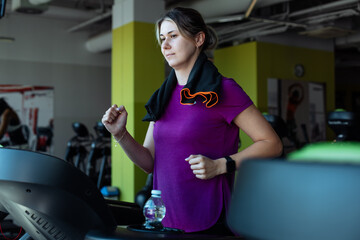 Overweight woman with pleasure run on treadmill at fitness gym with towel hanging around neck, side...