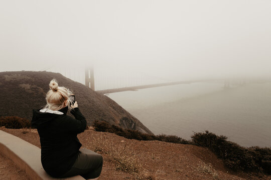 Woman and tourist taking a photo using mobile phone of Golden Gate Bridge and the landscape view of San Francisco Bay on a foggy and cloudy day in California, USA.