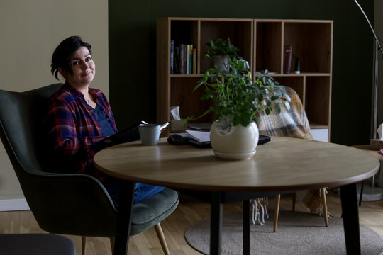 Happy Brunette Woman Relax With Cup Of Tea In Stylish Armchair At Table In Home Room. Reading Activity For Adult Female People, Enjoying Quiet Lifestyle Indoor. Resting Place In Modern Design