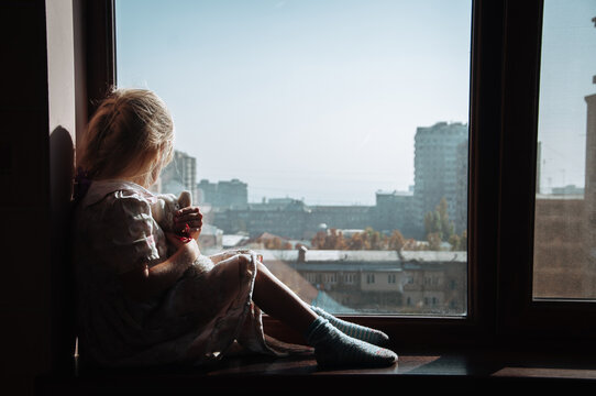 A girl with a toy is sitting on the windowsill. The child is bored.