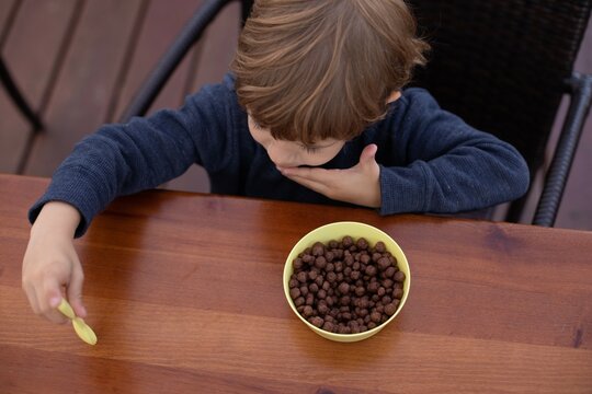 Little Boy Sitting On Chair At Brown Wooden Table In Kitchen And Having Breakfast With Chocolate Balls Top View. Kid Of Kindergarten Age Trifle Spoon In Hand. Ready Breakfast, Not Hungry Child