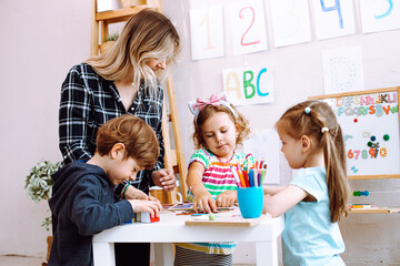 Group of kids playing together with educational toys in playroom of kindergarten. Young educator sitting with children at table on floor and help to play learning games. Lesson in daycare centre - obrazy, fototapety, plakaty