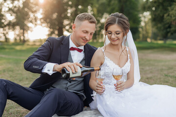 Portrait of wedding couple sitting on grass in park in summer. Young man groom pouring wine from...