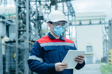 A masked power engineer during a pandemic inspects the modern equipment of an electrical substation before commissioning. Energy and industry. Scheduled repair of electrical equipment.