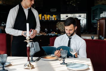 A young businessman in a fine restaurant examines the menu and makes an order to a young waiter in a stylish apron. Customer service. Table service in the restaurant.