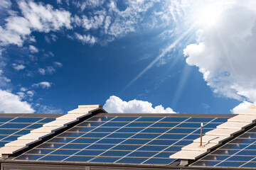Close-up of a roof with many solar panels against a clear blue sky with clouds, sunbeams and copy...