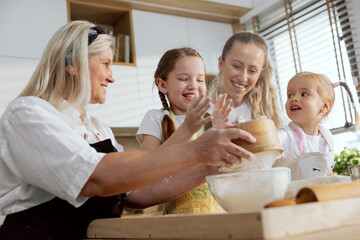 Fototapeta premium Grandmother mother and daughters cooking in the kitchen baking cookies. Granny sowing flour through sieve looking at preschooler grandchild talking. Girls having fun.