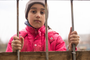 A little girl with a sad look behind a metal fence. Social problem of refugees and forced migrants