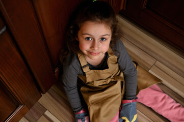 Overhead view of a Caucasian child, baby girl, cute little baker confectioner, chef pastry in a beige apron and mittens, sitting barefoot on the kitchen floor and cutely smiling looking at camera