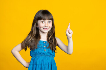 A little smiling girl of school age attracts attention by raising her index finger up and points to a place for an inscription for advertising and discounts. Isolated on yellow background