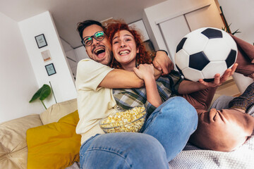 Happy young couple spending weekend together at home sitting on sofa watching soccer play live on...