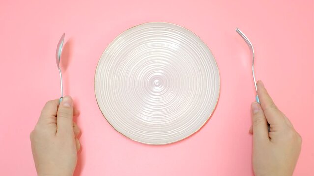 Female hands holding a fork and spoon next to an empty plate on a pink background close-up top view.