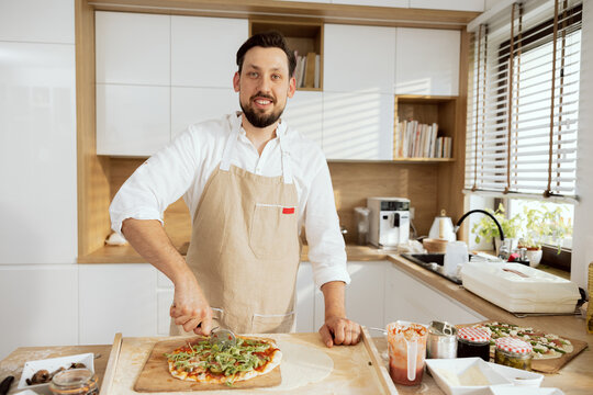 Cheerful man husband father in aprons standing at wooden table in modern kitchen cutting homemade pizza. Making different taty pizza at home preparing lunch for children.