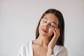 Close up tender young caucasian woman gently touching her face with closed eyes at home. Women's brown hair, wears white clothes. Skin care concept
