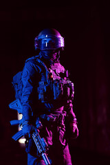 Army soldier in Combat Uniforms with an assault rifle and combat helmet night mission dark background. Blue and purple gel light effect. 