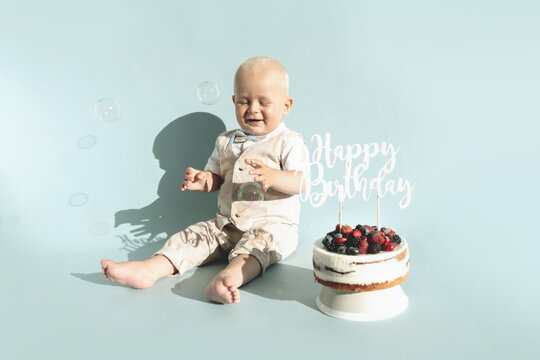 Happy, surprised one year baby boy eats sweets. Toddler boy and first healthy homemade birthday cake with berries on blue background. Copy space. Child's emotions. Party celebration, greeting card