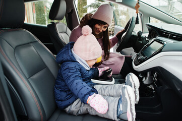 Young mother with baby girl daughter sit in electric car in the yard of her house at winter.