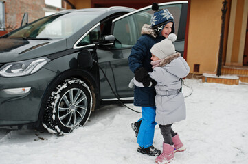 Brother with sister play against charging electric car in the yard of house at winter.