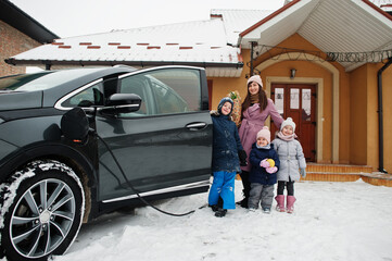 Young woman with kids hold eco bags and charging electric car in the yard of her house .