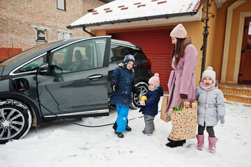 Young woman with kids hold eco bags and charging electric car in the yard of her house .