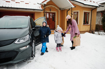 Young woman with kids hold eco bags and charging electric car in the yard of her house .