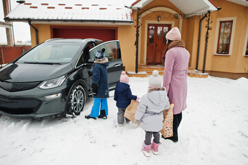 Young woman with kids hold eco bags and charging electric car in the yard of her house .