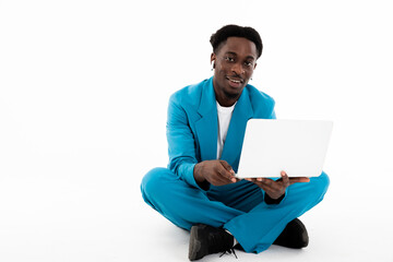 Photo of handsome african american man sitting on white floor in lotos pose holding laptop in hands working online posing for camera.