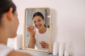 Positive young caucasian woman rubs face with cucumber slices and smiling at camera home. Brown hair girl has lot of skin care products on table. Concept spa morning, beauty.