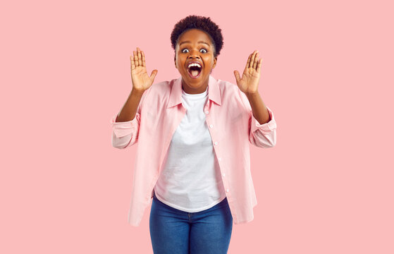 Overjoyed attractive African American woman screams with wonder or joy experiences WoW emotions and raises hands when he sees unexpected surprise or meets friend stands on pink background
