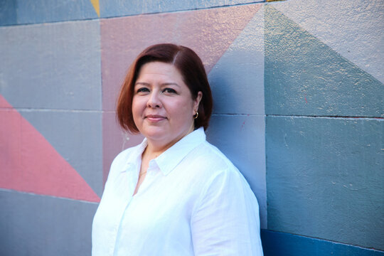 Smiling middle aged woman with short hair wearing white shirt leaning on a coloured wall