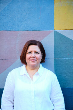 middle aged woman with short hair wearing white shirt leaning on a coloured wall