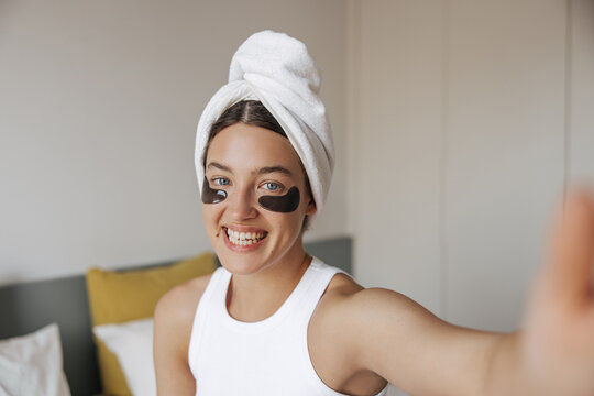 Happy young caucasian woman with towel on head and eyes patches takes selfie on smartphone. Brunette hair female sitting bed in her bedroom. Concept selfie, positive emotions.