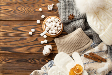 Fototapeta na wymiar Winter concept. Top view photo of cup of cocoa with marshmallow on rattan serving mat jumper bobble hat scarf mittens cotton flower cinnamon sticks dried orange slices anise on wooden desk background