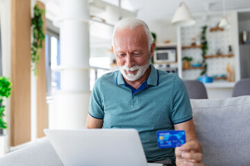 Mature man using laptop, holding plastic credit or debit card, senior grey haired customer making secure internet payment, shopping or browsing online banking service, entering information