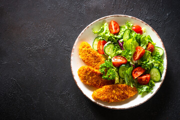 Healthy green salad with chicken nuggets in craft plate at black background. Top view image with copy space.