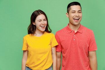 Young cheerful smiling happy fun couple two friends family man woman wear basic t-shirts together looking camera wink blink eye isolated on pastel plain light green color background studio portrait.