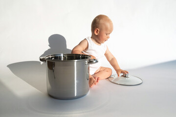 Little cute baby chef sitting near big cooking pot with kitchen, utensils, accessories on white background. Child cook, food boy. Copy space
