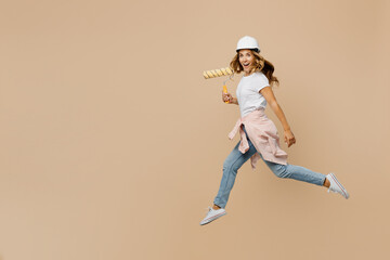 Fototapeta na wymiar Full body young employee laborer handyman woman in white t-shirt helmet jump high hold wall paint roller isolated on plain beige background Instruments accessories renovation room Repair home concept