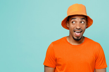 Young surprised happy man of African American ethnicity 20s wear orange t-shirt hat look aside on workspace area mock up isolated on plain pastel light blue cyan background. People lifestyle concept.