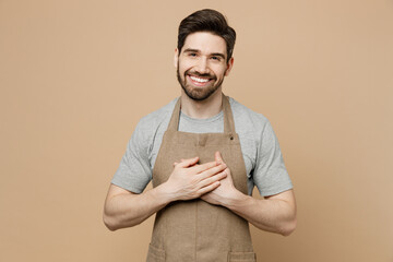 Young happy fun professional man barista barman employee wear brown apron work in coffee shop put folded hands on heart isolated on plain pastel light beige background. Small business startup concept.