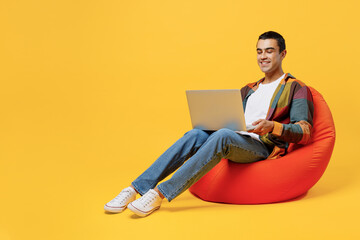 Full body young middle eastern man he wear casual shirt white t-shirt sit in bag chair IT woman hold use work on laptop pc computer isolated on plain yellow background studio People lifestyle concept.