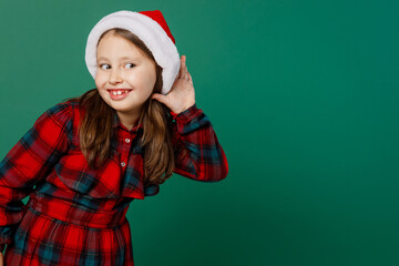 Merry curious little child kid girl 6-7 years old wear red dress Christmas hat posing try to hear you overhear listening intently isolated on plain dark green background. Happy New Year 2023 concept.