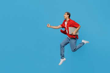 Fototapeta na wymiar Full body side view young IT man of African American ethnicity wear red shirt hold use work on laptop pc computer jump high run isolated on plain pastel blue cyan background. People lifestyle concept.