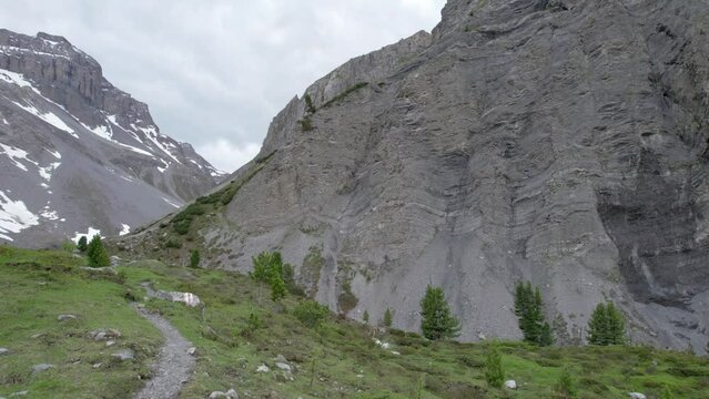 Aerial drone footage panning through a glacial mountain landscape with patches of snow, isolated trees a remote alpine hiking trail in Switzerland.