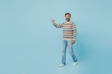 Full body young smiling happy cheerful man 30s he wears sweater walking going point index finger aside on workspace area isolated on plain pastel light blue cyan background. People lifestyle concept.