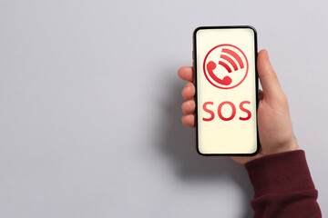 Man holding smartphone with emergency call SOS on screen on light grey background, space for text