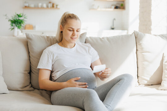 Happy pregnant woman looking at ultrasound scan of baby on sofa, light kitchen background
